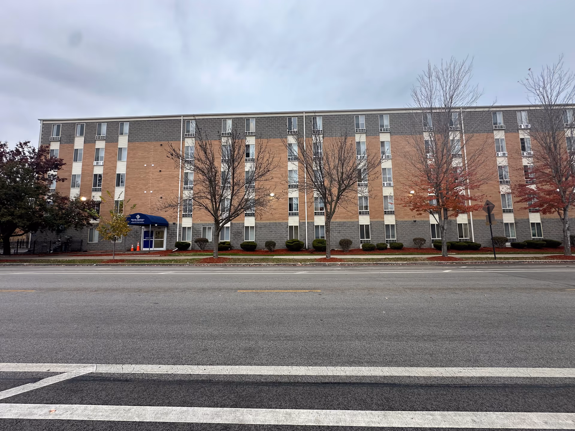Exterior front view of a multi-story brick and concrete building with several windows, a blue awning entrance labeled Grand Regency of Jackson Park Supportive Living, and leafless trees along the sidewalk under a cloudy sky.