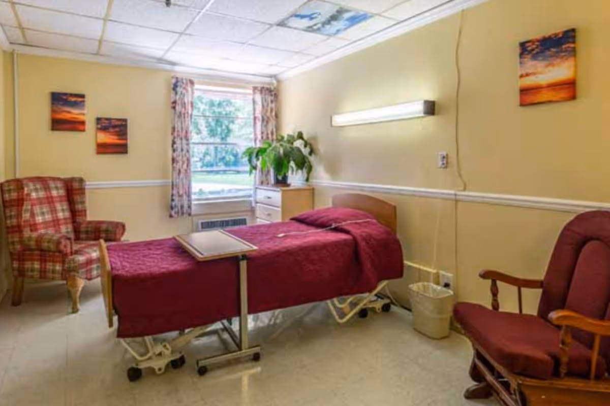 Senior living bedroom with maroon bed, window, plant, dresser, and chairs.