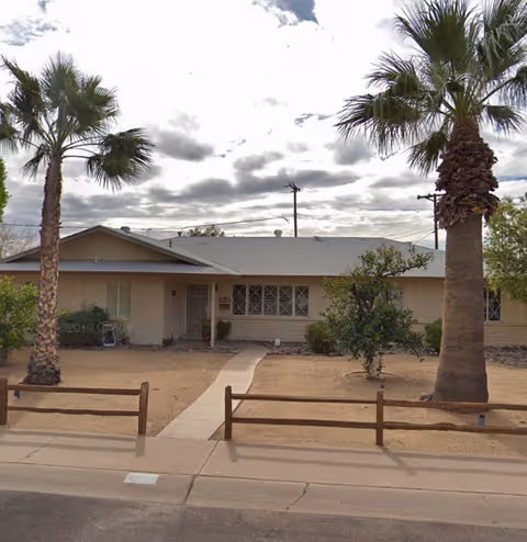 Single-story building with a light-colored exterior, a pathway leading to the front door, two tall palm trees, and a small fenced area in front. The sky is cloudy.