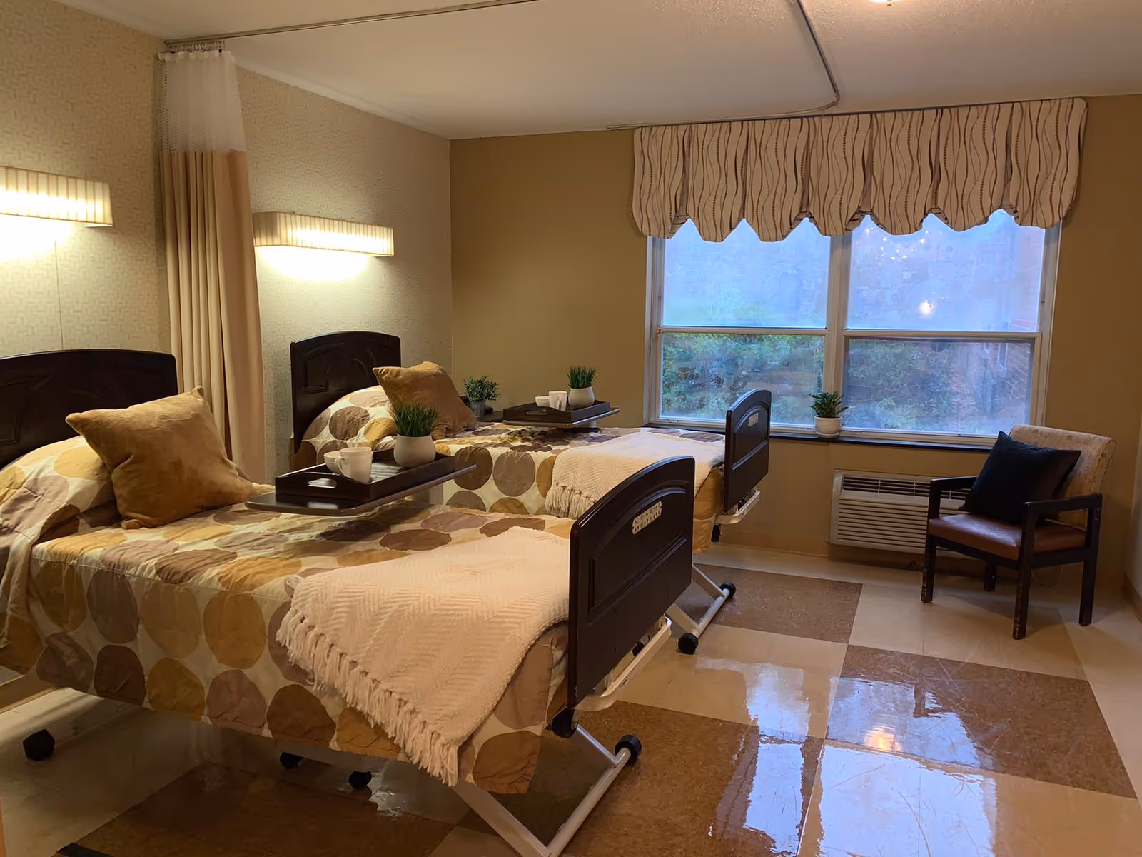 A well-lit room with two hospital-style beds, each with a beige and brown patterned bedspread and a beige throw blanket. Each bed has a tray with a white cup and a small green plant. There is a window with a beige valance and a chair with a dark cushion near the window. The floor has a checkered pattern with brown and beige tiles.
