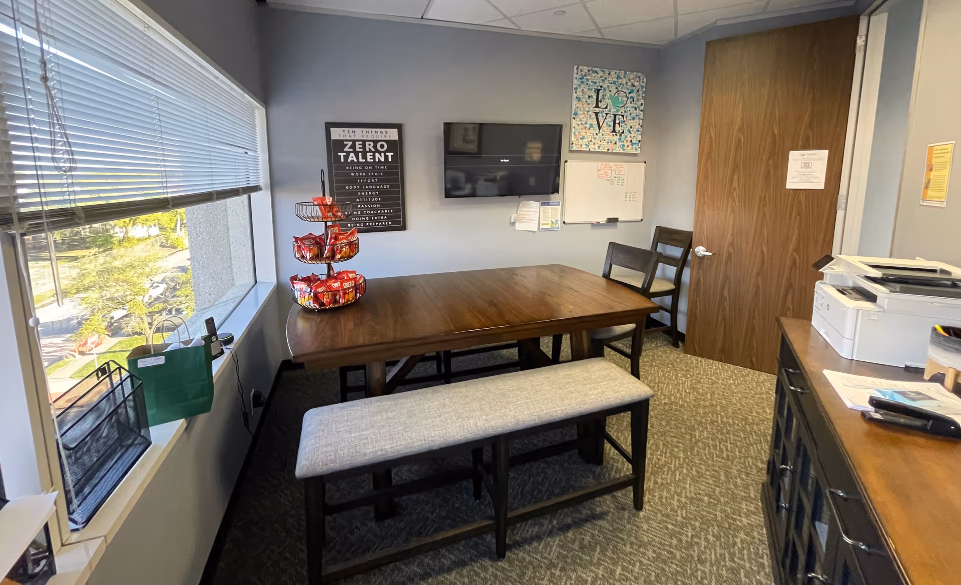 Small interior meeting room with a wooden table, bench seating, window, decor, and a printer.