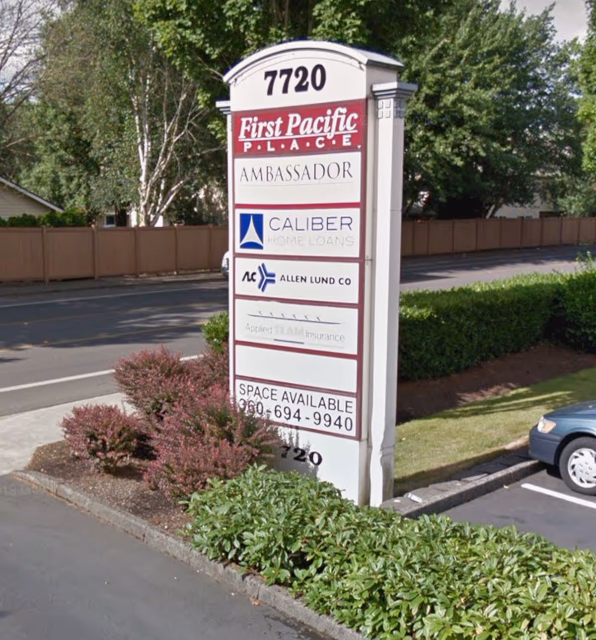 Outdoor signboard listing multiple businesses beside shrubbery, near a road.
