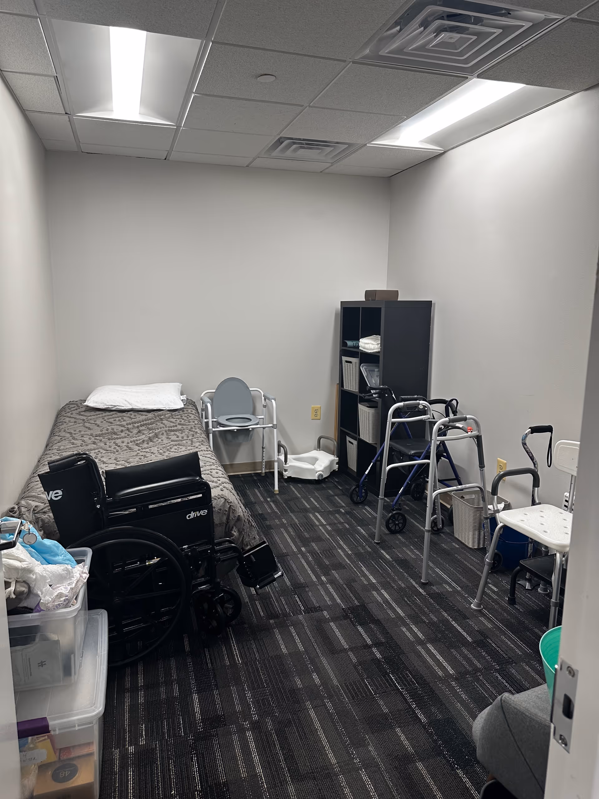 Small care room with bed, wheelchair, walker, and shelves in a neutral interior.