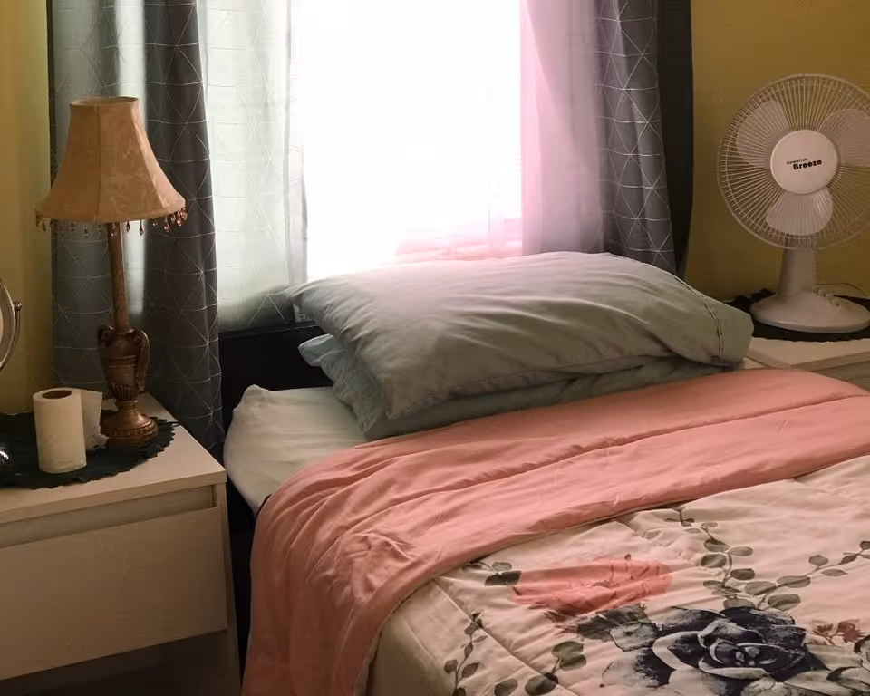 A tidy bedroom with pink floral bedspread, pillows, lamp, tissue, and a desk fan.