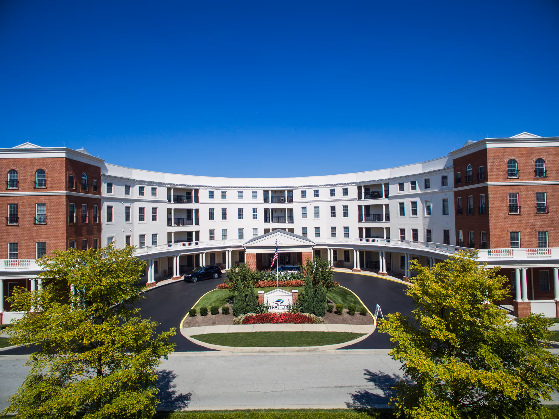 Curved four-story red-brick and white-faced senior living building with a circular driveway and landscaped entrance under a clear blue sky.