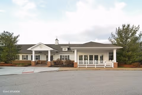 Front exterior view of a single-story senior living facility building with white siding, a covered entrance supported by columns, and a small fenced patio area. There are trees and landscaping around the building under a partly cloudy sky.