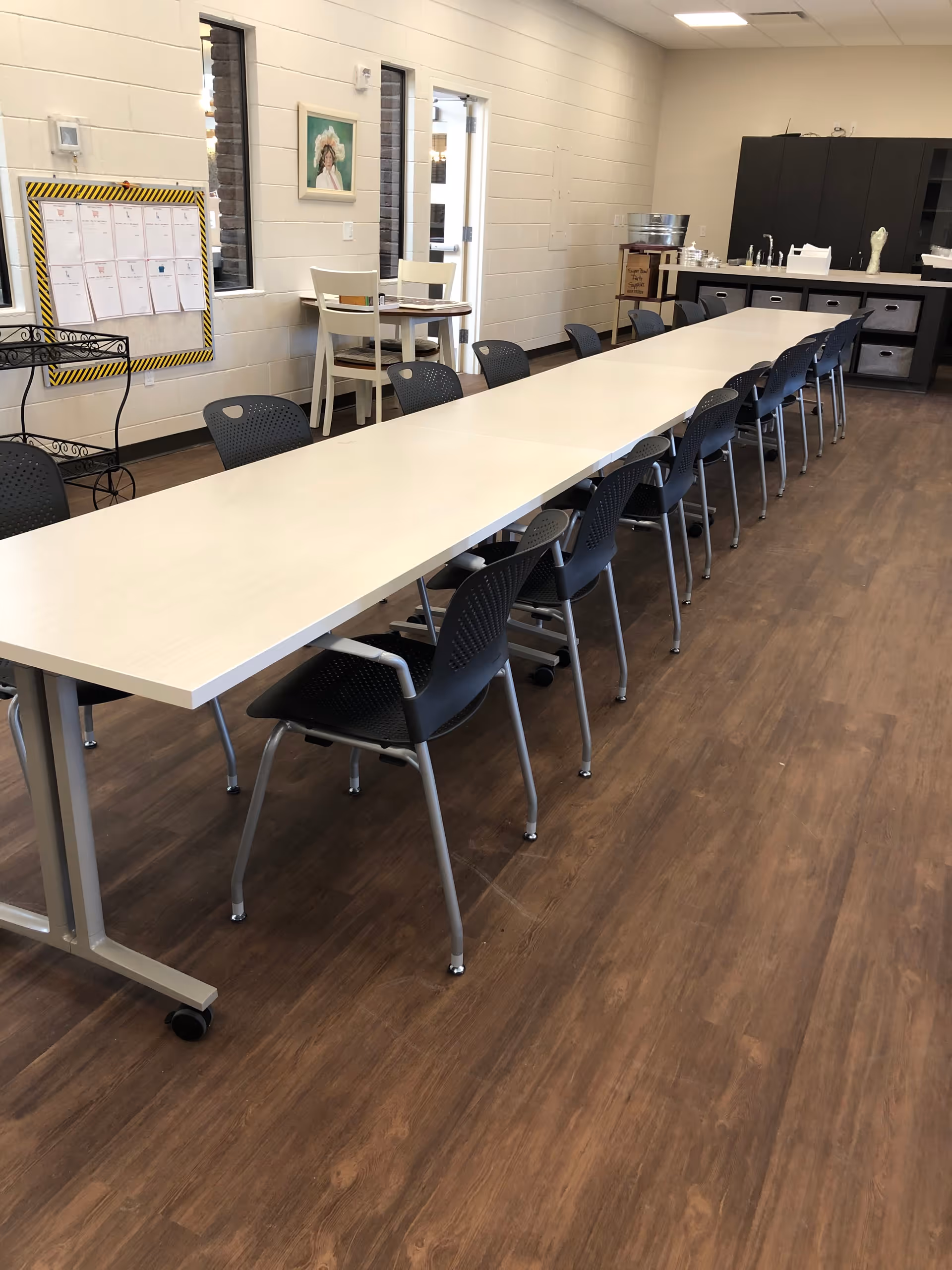 A long white table with multiple black chairs arranged on both sides in a room with wooden flooring. In the background, there is a small round table with two chairs near a window and a door. The room has white walls, a bulletin board, a painting, and a counter with cabinets and various items on top.