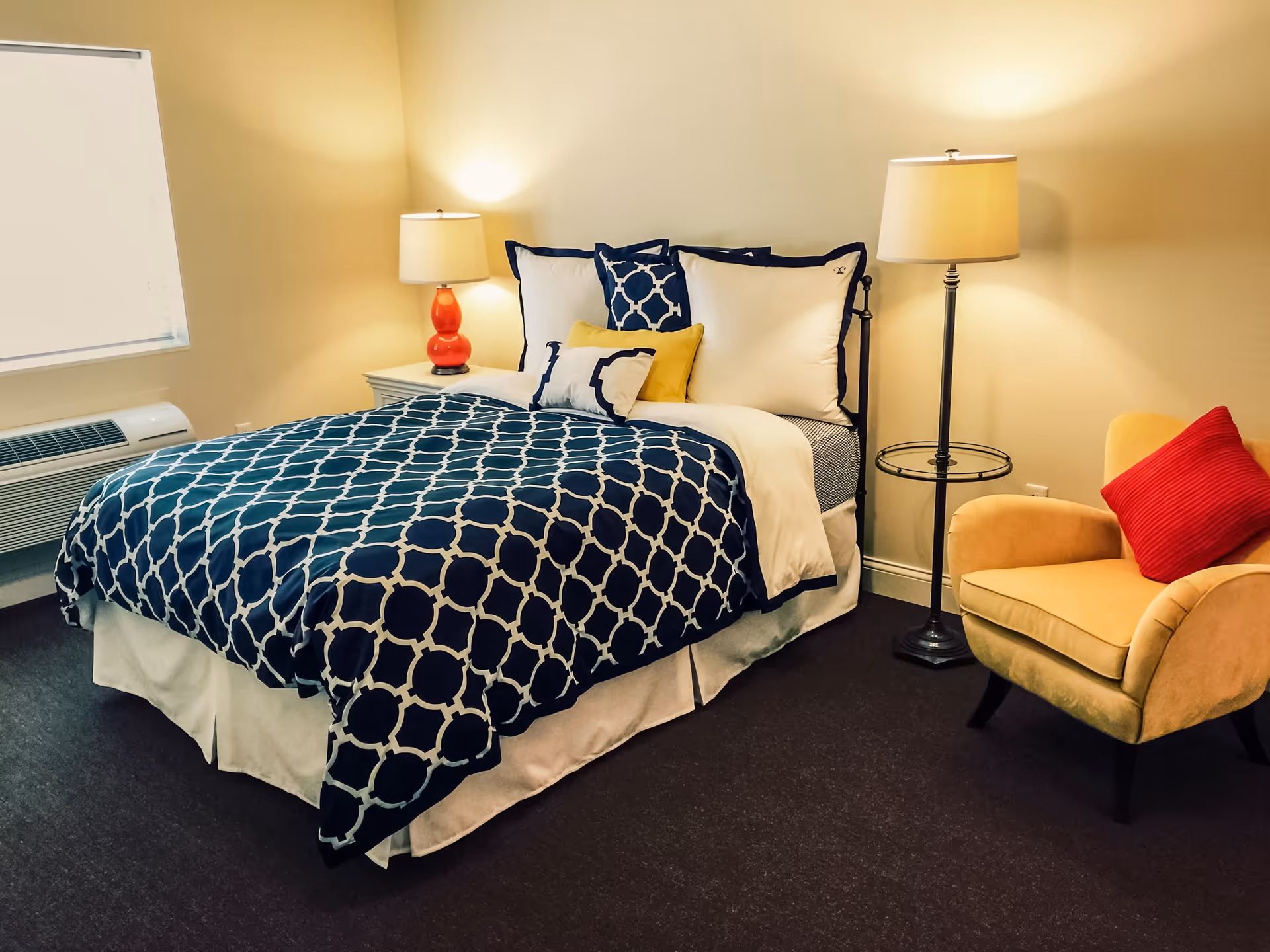A neatly made bed with a blue patterned comforter, two bedside lamps, and a yellow armchair with a red pillow in a bedroom.