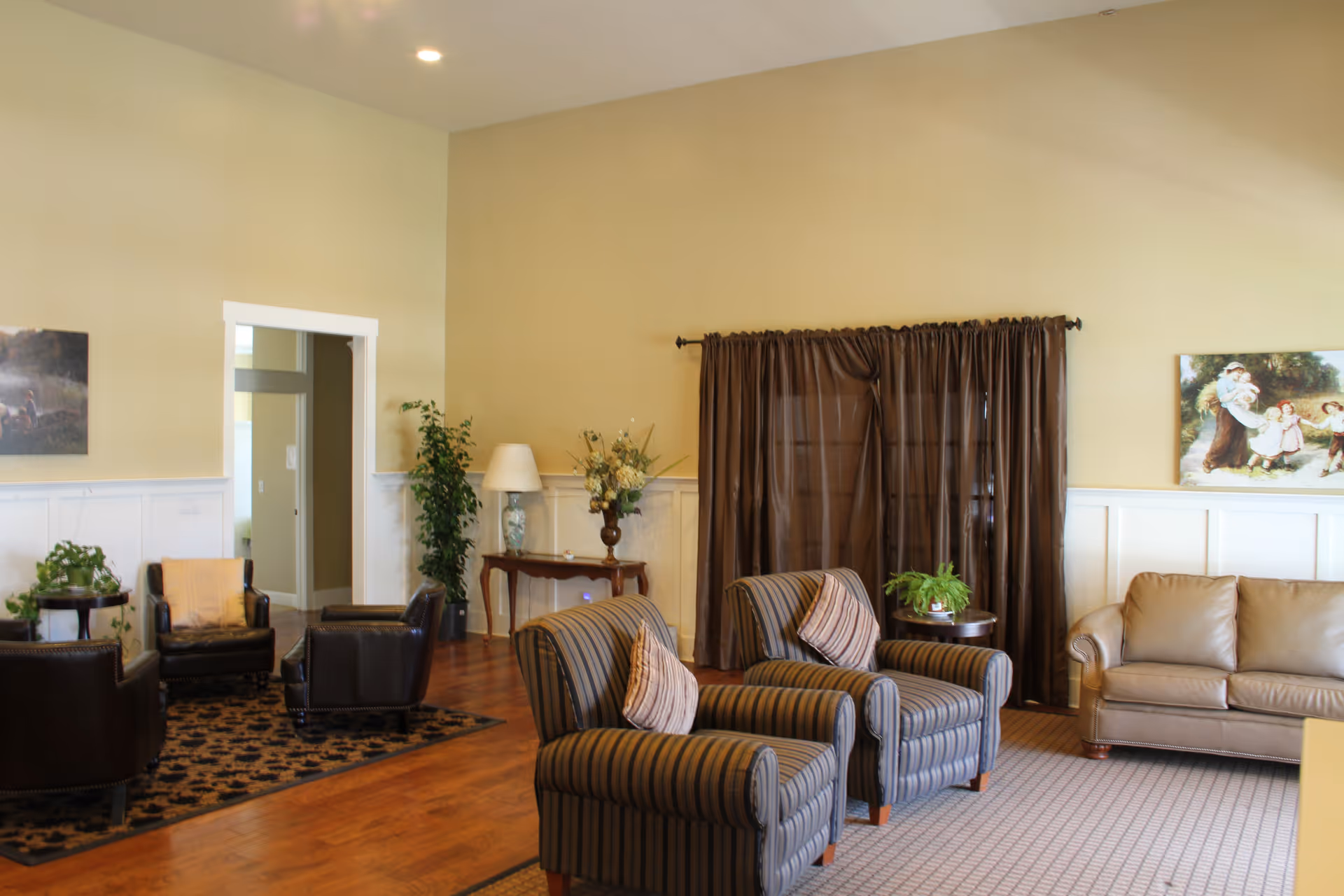A spacious senior living lounge with striped chairs, a sofa, plants, and brown curtains.