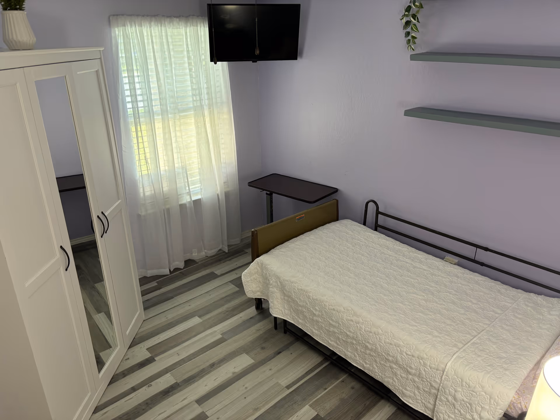 A furnished bedroom in a senior living facility with a bed, wardrobe, table, and sheer curtains.
