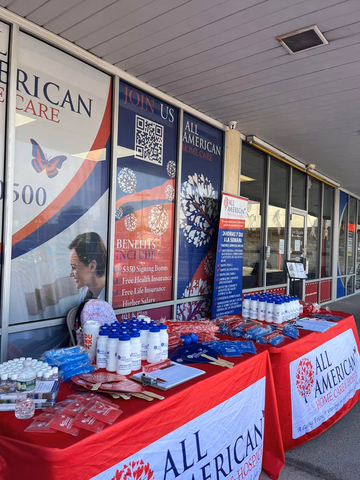 Outdoor promotional booth in front of All American Home Care banners and tables.