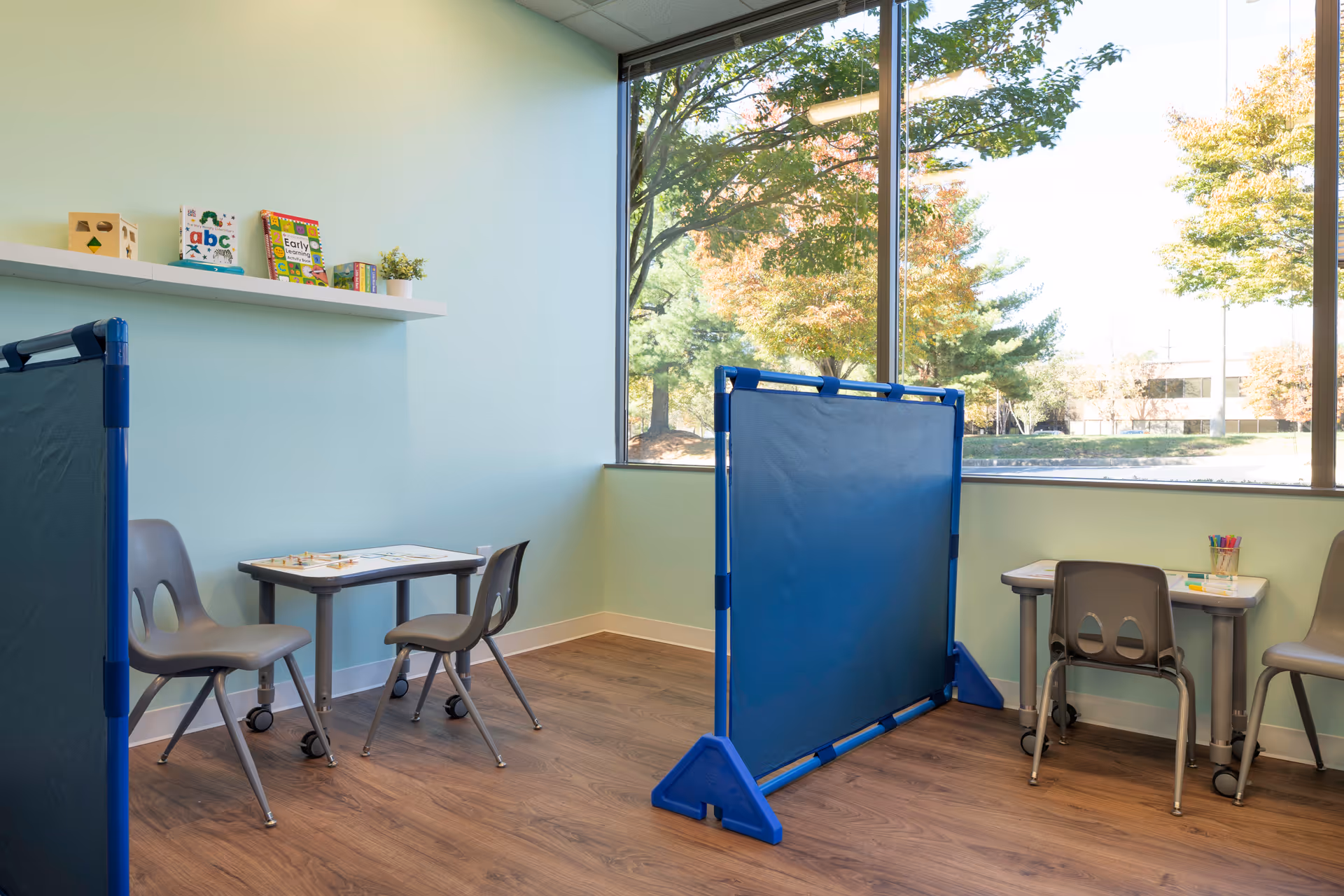 Bright children's activity room with small tables, chairs, and a blue divider panel.
