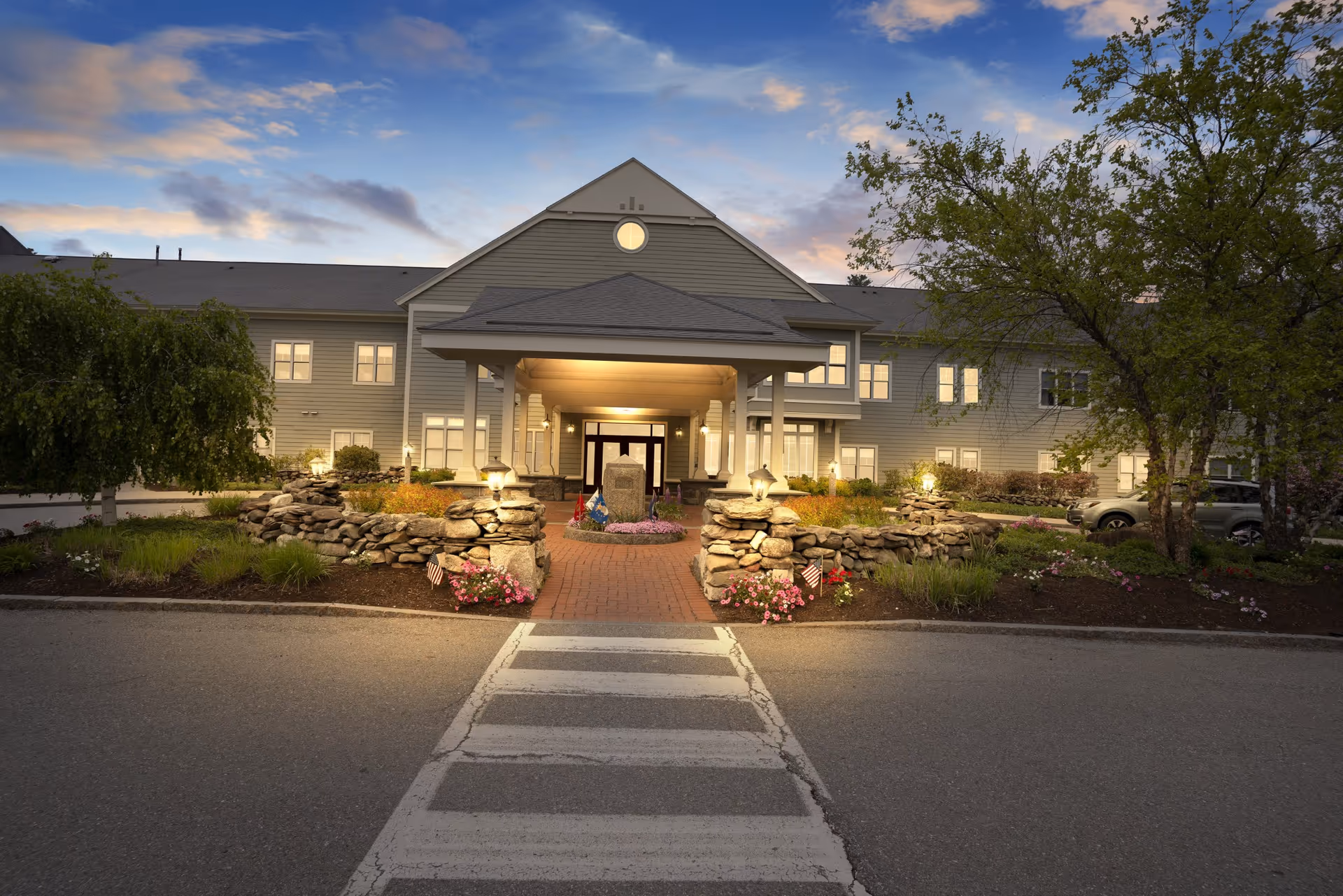 Front exterior view of Dirigo Pines facility at dusk with a covered entrance, stone walls, landscaped garden beds, and a crosswalk leading to the entrance.