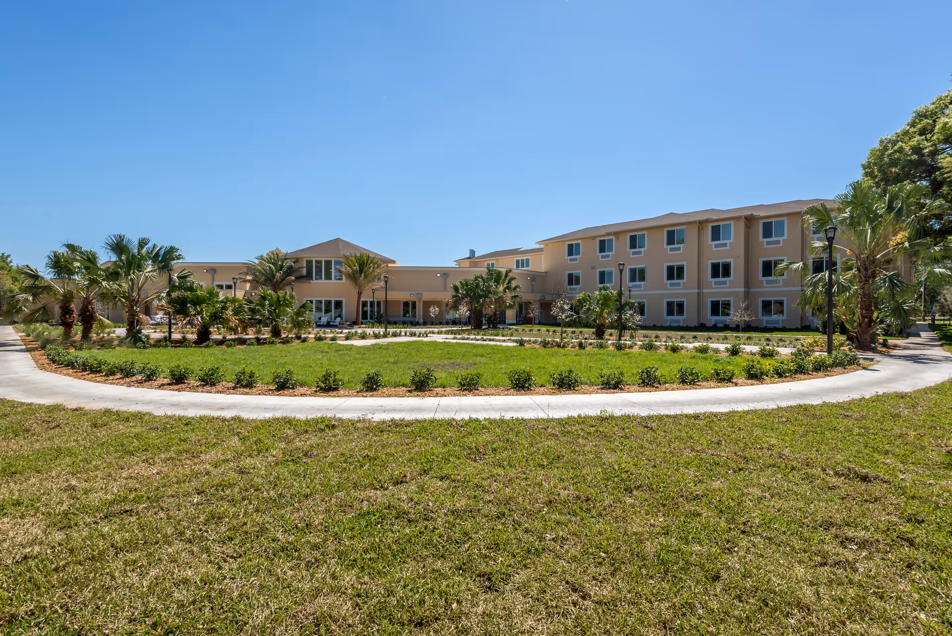Front view of a three-story beige building with landscaped lawn, palm trees, and a circular driveway under a clear blue sky.
