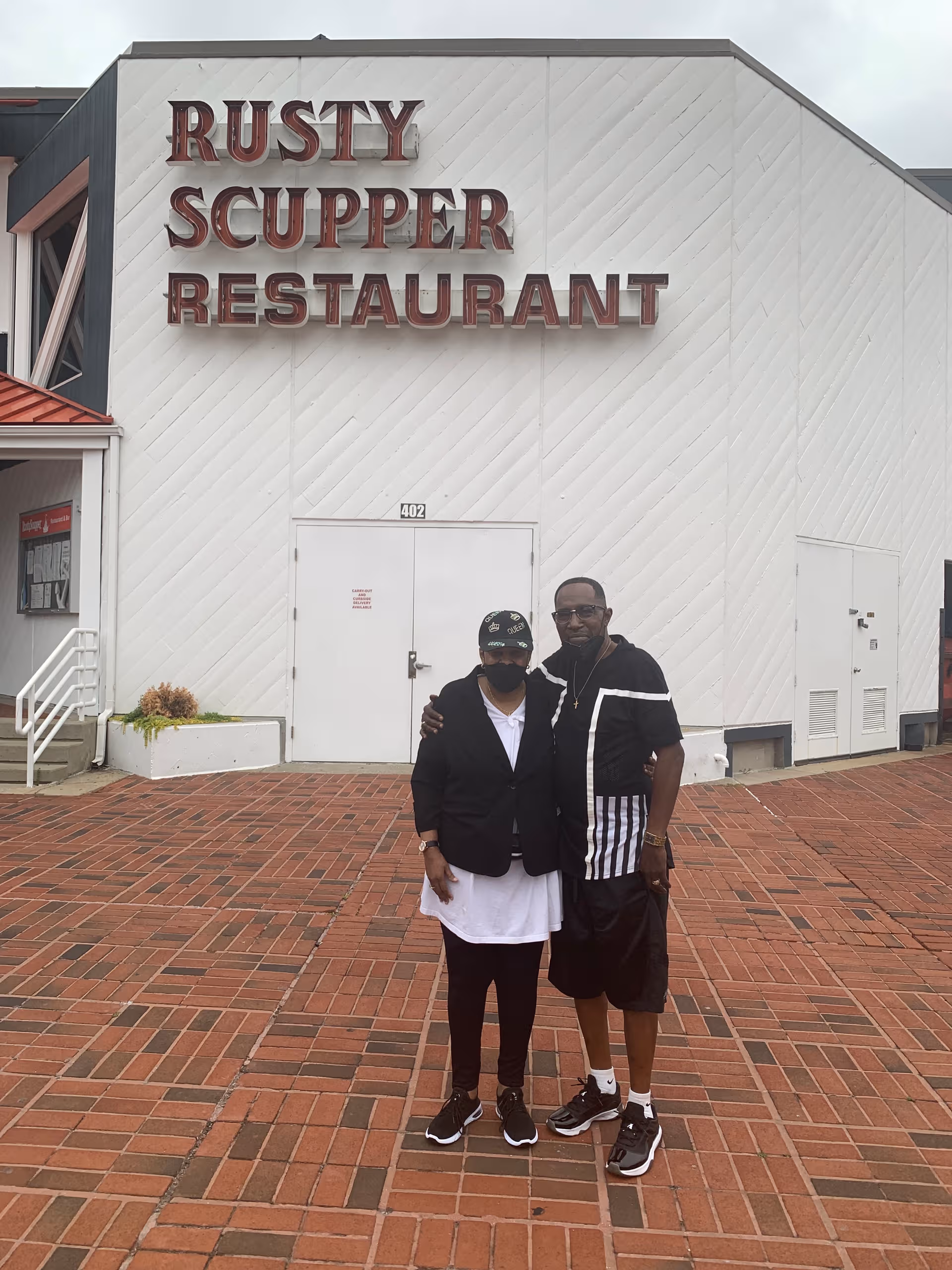 Two people stand in front of a white building with a Rusty Scupper Restaurant sign.