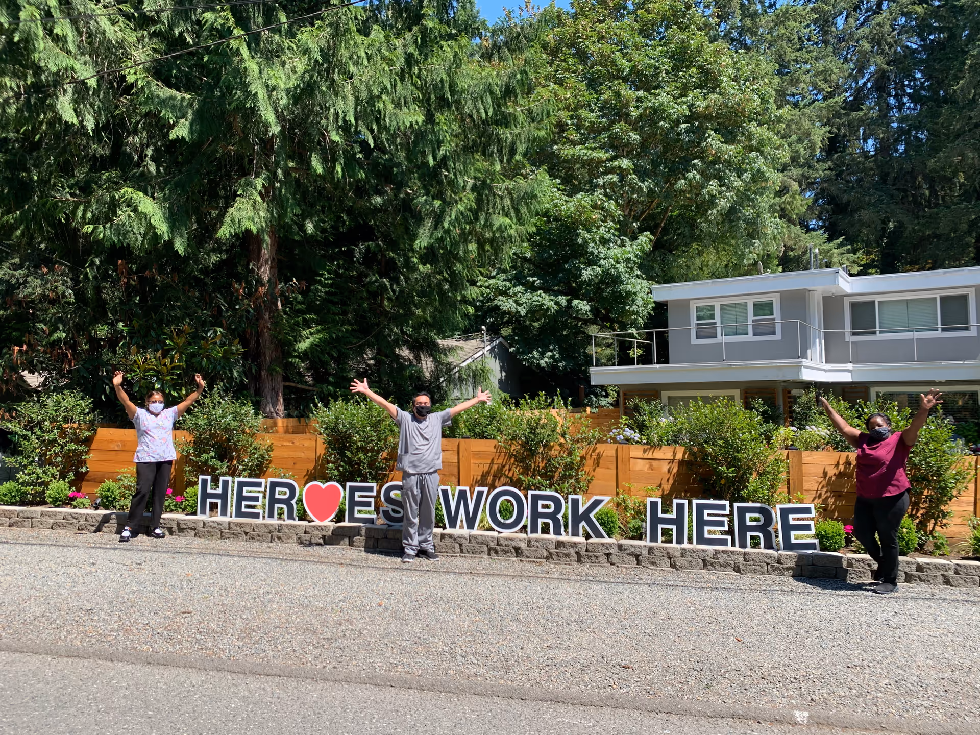 Three people wearing masks standing outside in front of a wooden fence and greenery, with a large sign that reads 'HEROES WORK HERE' with a red heart symbol replacing the letter O in HEROES. They have their arms raised in celebration.