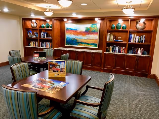 A cozy interior room with two wooden tables surrounded by striped upholstered chairs. Behind the tables is a large wooden built-in bookshelf filled with books, decorative vases, and a colorful abstract painting in the center. The room is softly lit with ceiling lights and has a carpeted floor.