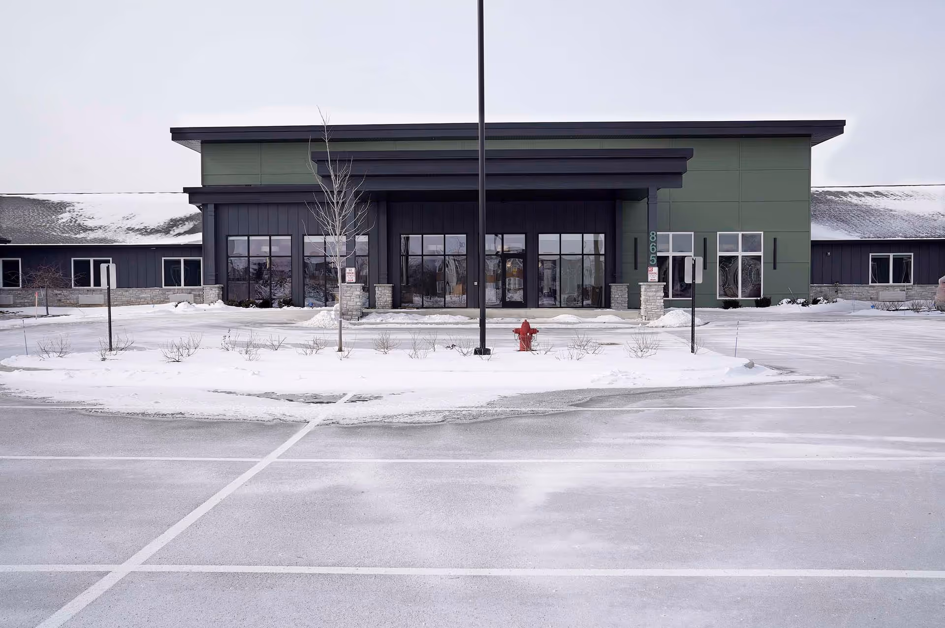 Snow-covered parking lot in front of a modern facility entrance with glass doors.