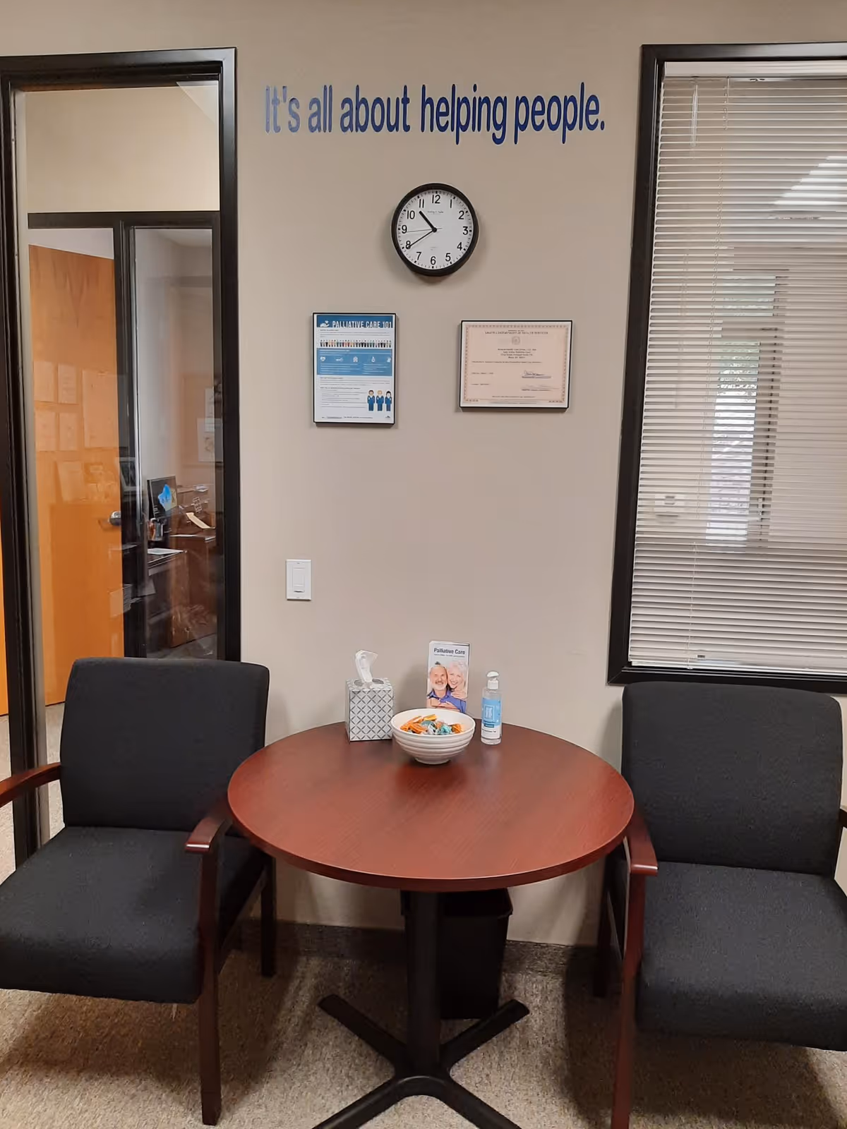 A small indoor waiting area with a round table, two chairs, a clock, and wall signage.