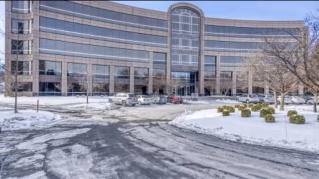 Snow-covered driveway and parking lot in front of a curved glass-faced building, likely a senior facility.