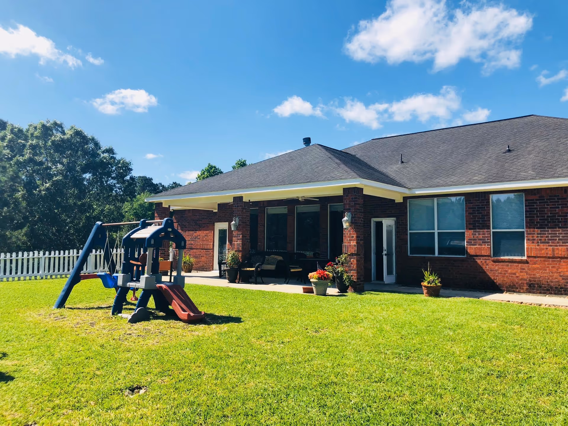 A sunny outdoor area of a brick building with a covered patio furnished with chairs and potted plants. In the foreground, there is a small children's playset with a slide and swings on a grassy lawn, surrounded by a white picket fence and trees in the background under a blue sky with some clouds.