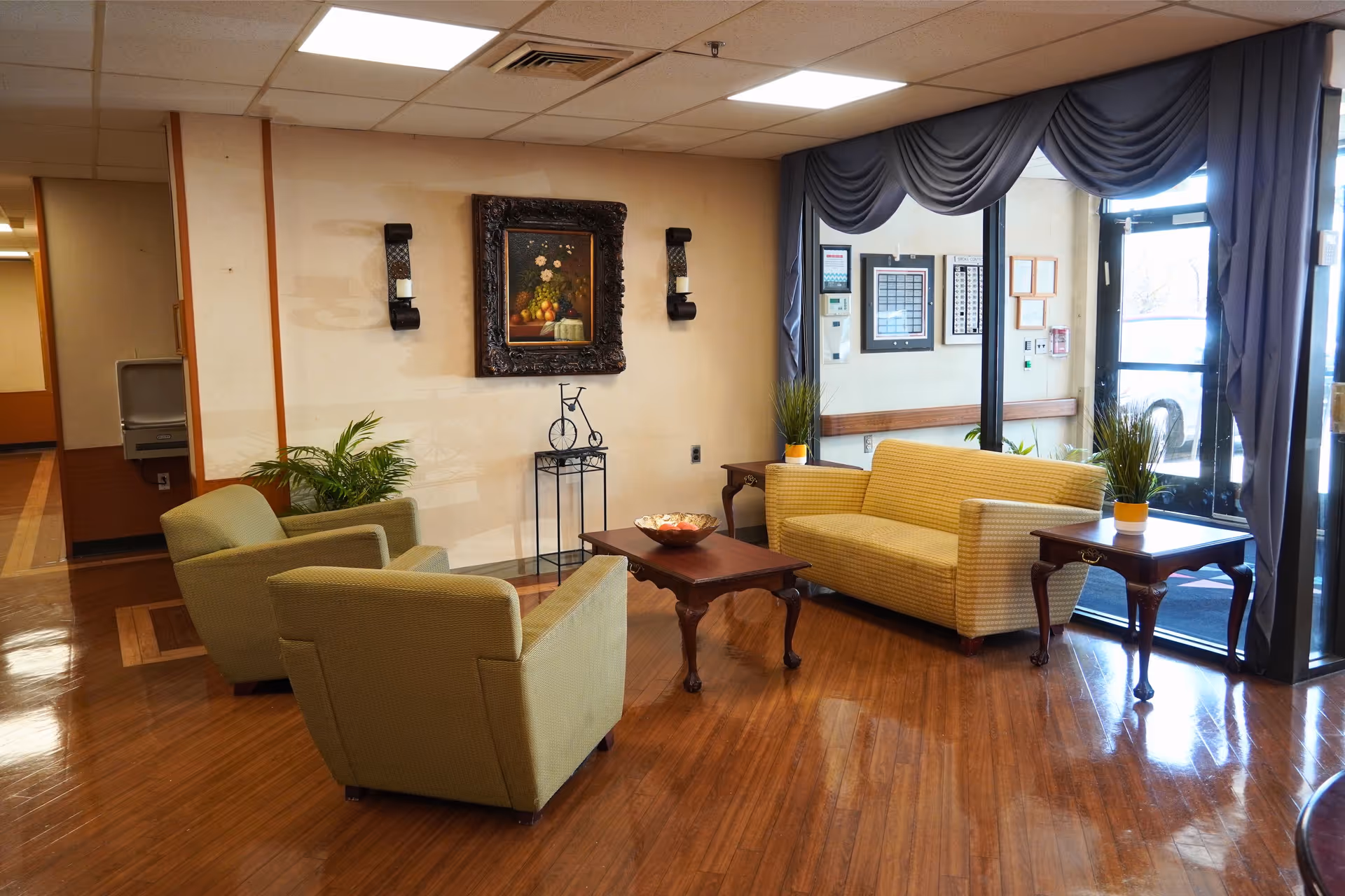 A cozy sitting area in a senior living facility with two green armchairs and a yellow sofa arranged around a wooden coffee table. The room has polished wooden floors, a framed painting on the wall, two wall sconces, and large windows with blue drapes letting in natural light. There are small plants on side tables and a water fountain visible in the hallway.