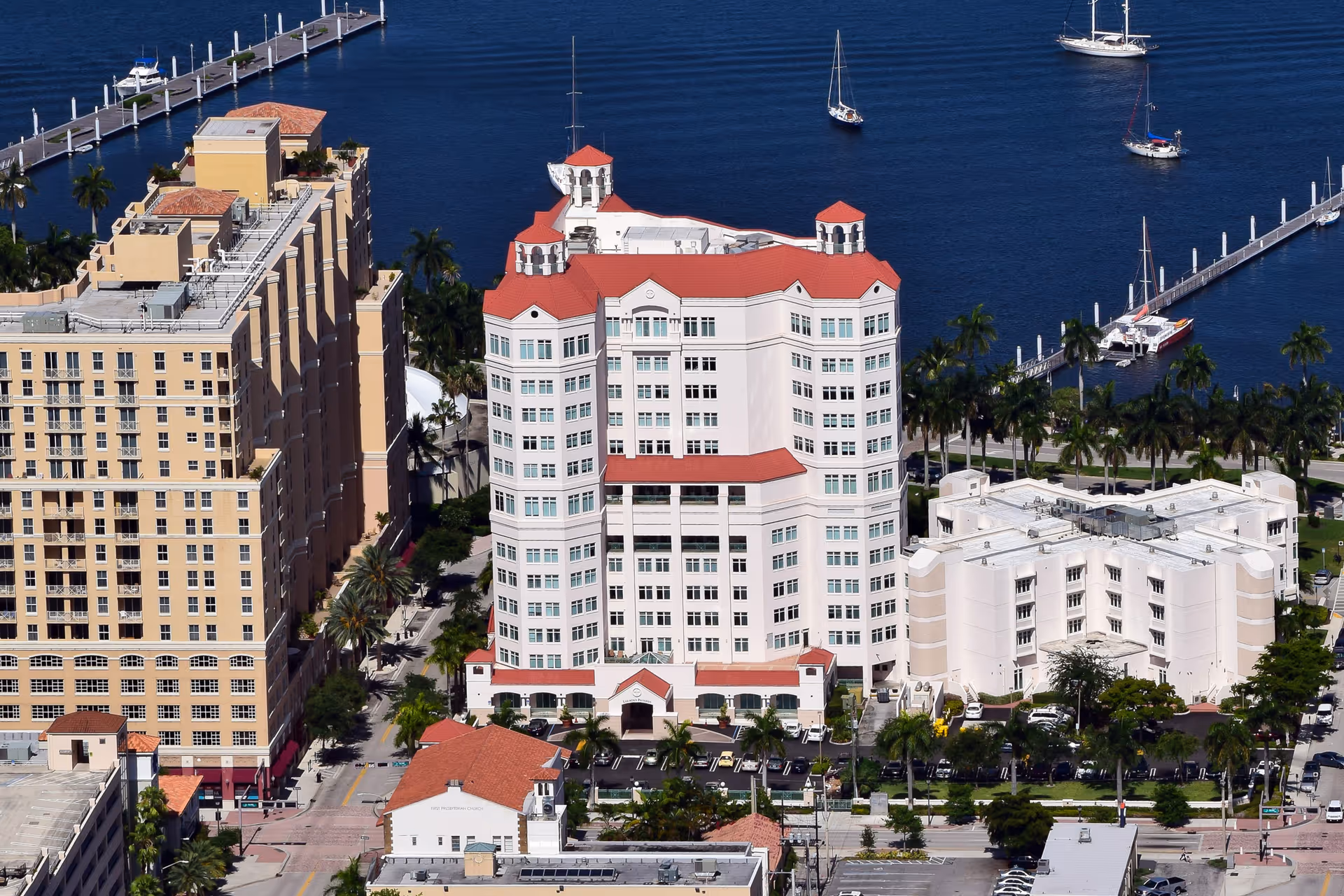 Aerial view of a multi-story white building with red roofs by a marina and sailboats.