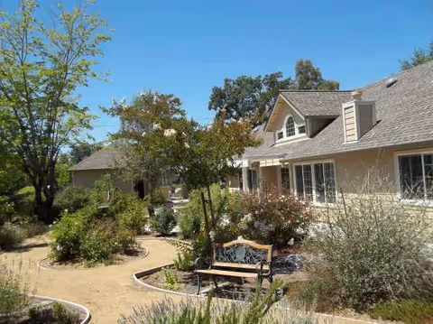 Senior living facility exterior with beige building, lush garden, and wooden bench along a winding path.