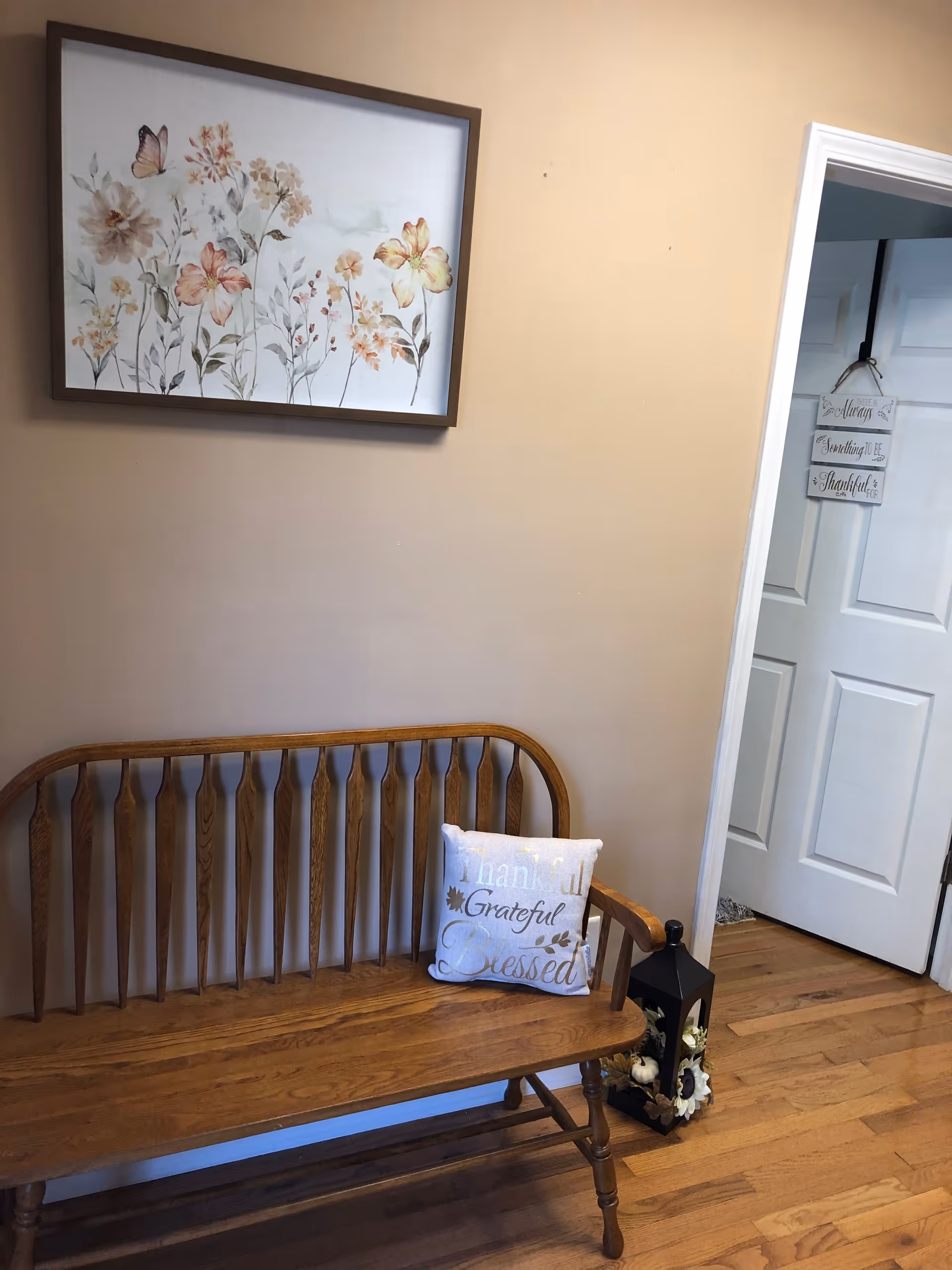 Framed floral wall art above a wooden bench with pillow and lantern beside a doorway.