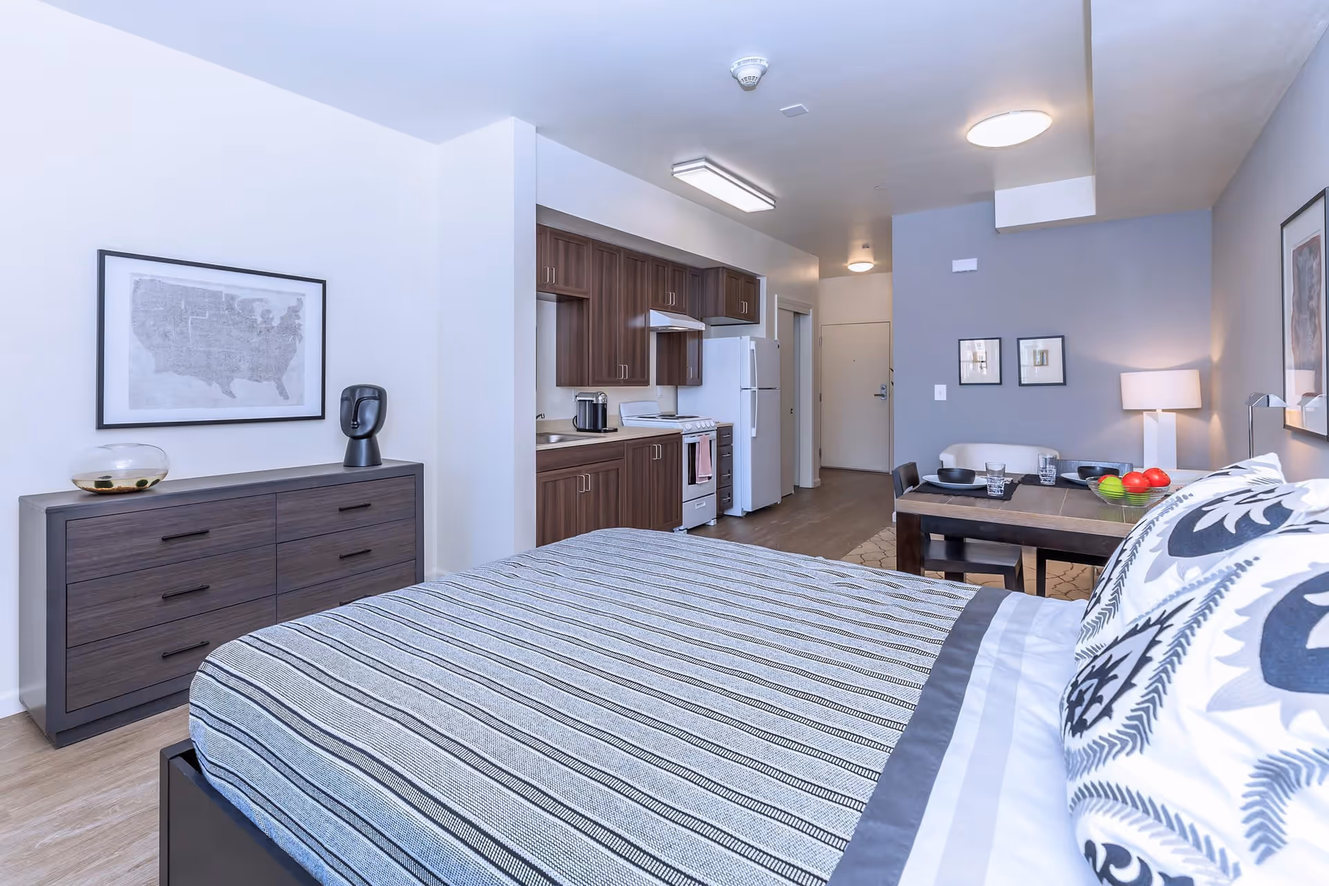 Bright open-plan studio interior with a bed in the foreground, dresser, kitchenette and dining table.