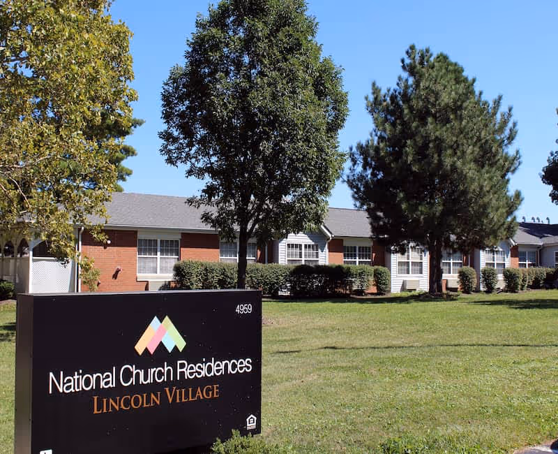 Front exterior of a single-story senior living building with trees, lawn, and a sign for National Church Residences Lincoln Village.
