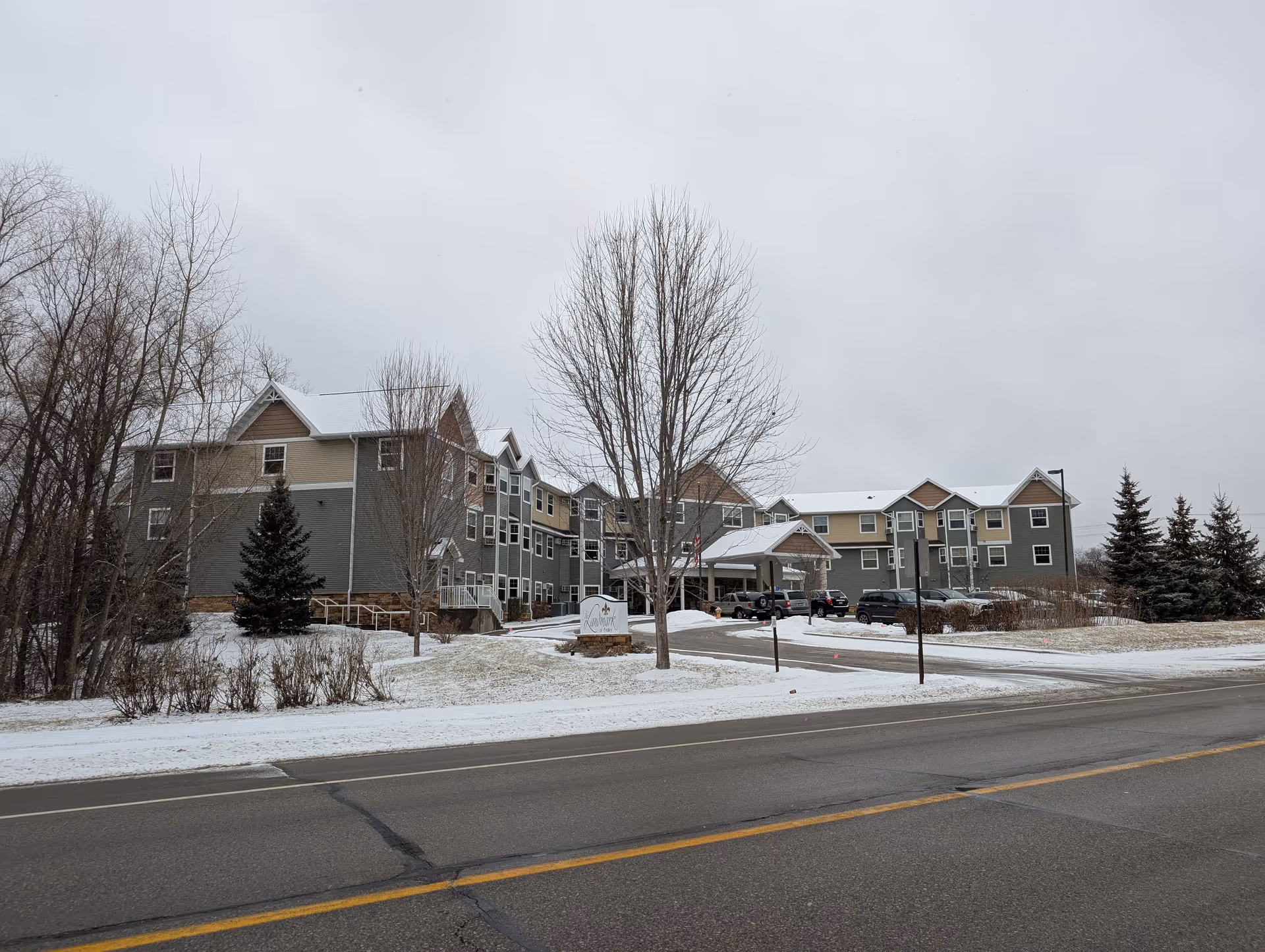 Front exterior of a three-story senior living building with a covered entrance, parked cars, bare trees, and light snow on the ground.