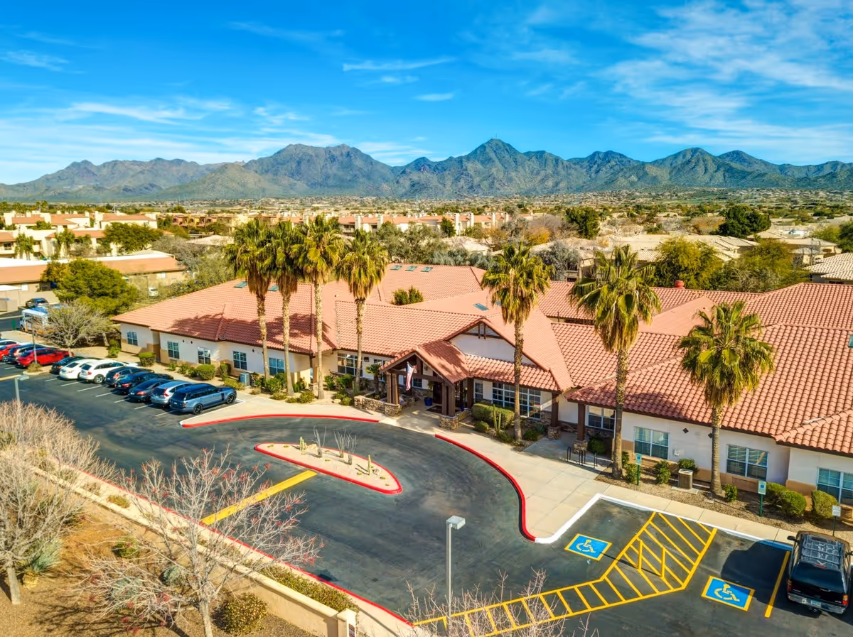 Aerial view of a senior living facility with red tile roofs, palm trees, a parking lot, and mountains in the background.