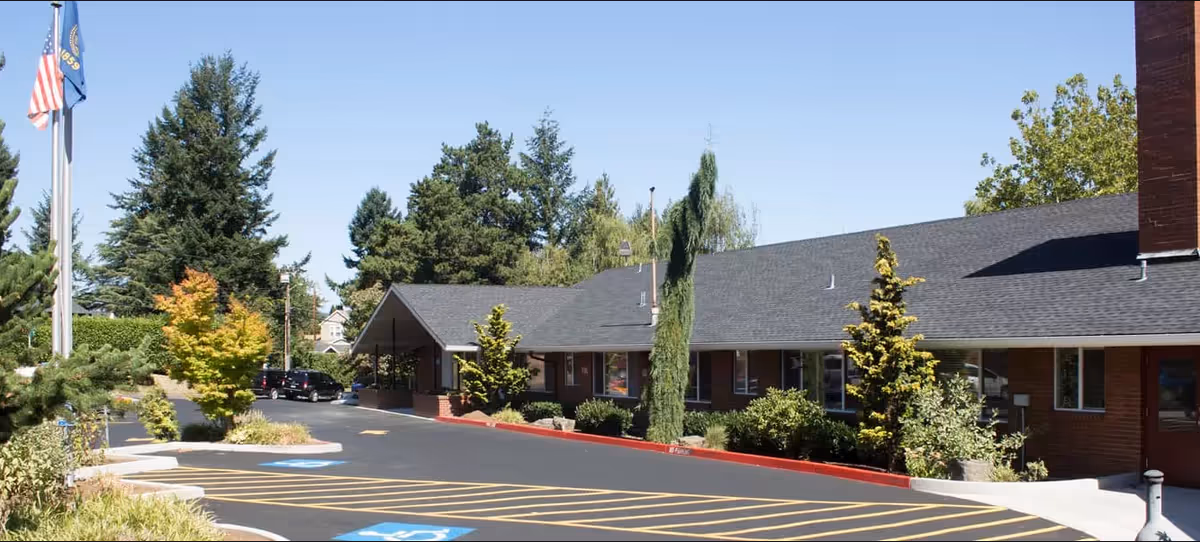 Exterior view of Avamere Crestview of Portland with a low-slung brick building, parking lot, and flagpoles.