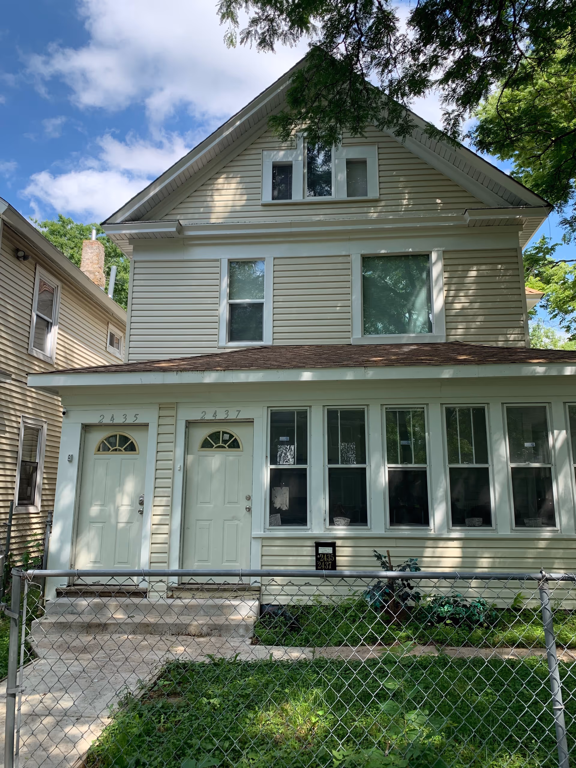 Two-story beige house with front doors, large windows, and a chain-link fence.