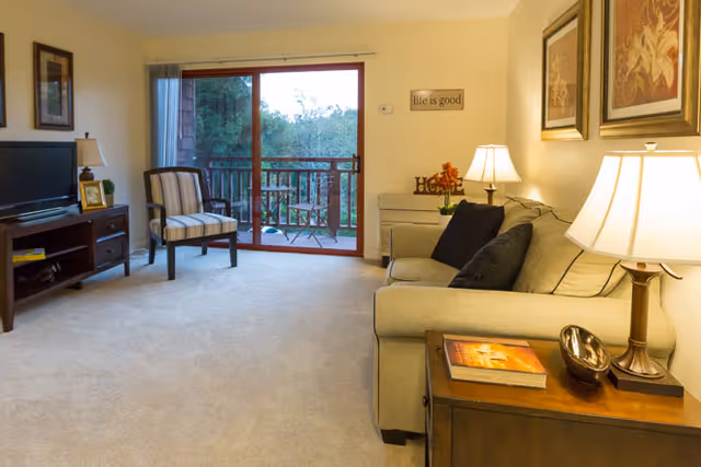 A cozy living room with a beige sofa adorned with dark cushions, a wooden side table with a lamp and decorative items, a striped armchair near a sliding glass door leading to a balcony with outdoor seating, a TV on a wooden stand, and framed artwork on the walls.