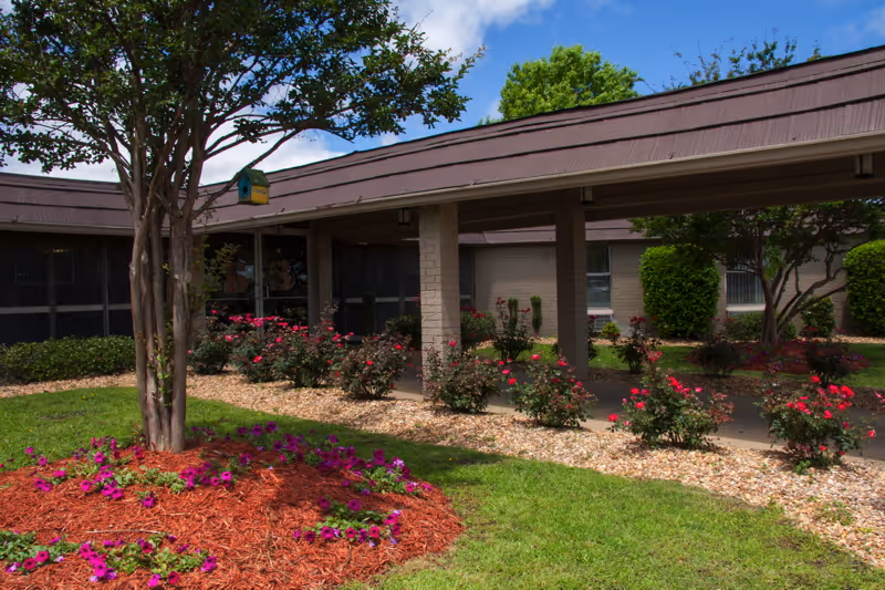 Outdoor garden area at a senior living facility with a covered walkway supported by columns, flower beds with red and purple flowers, green grass, and trees under a partly cloudy blue sky.