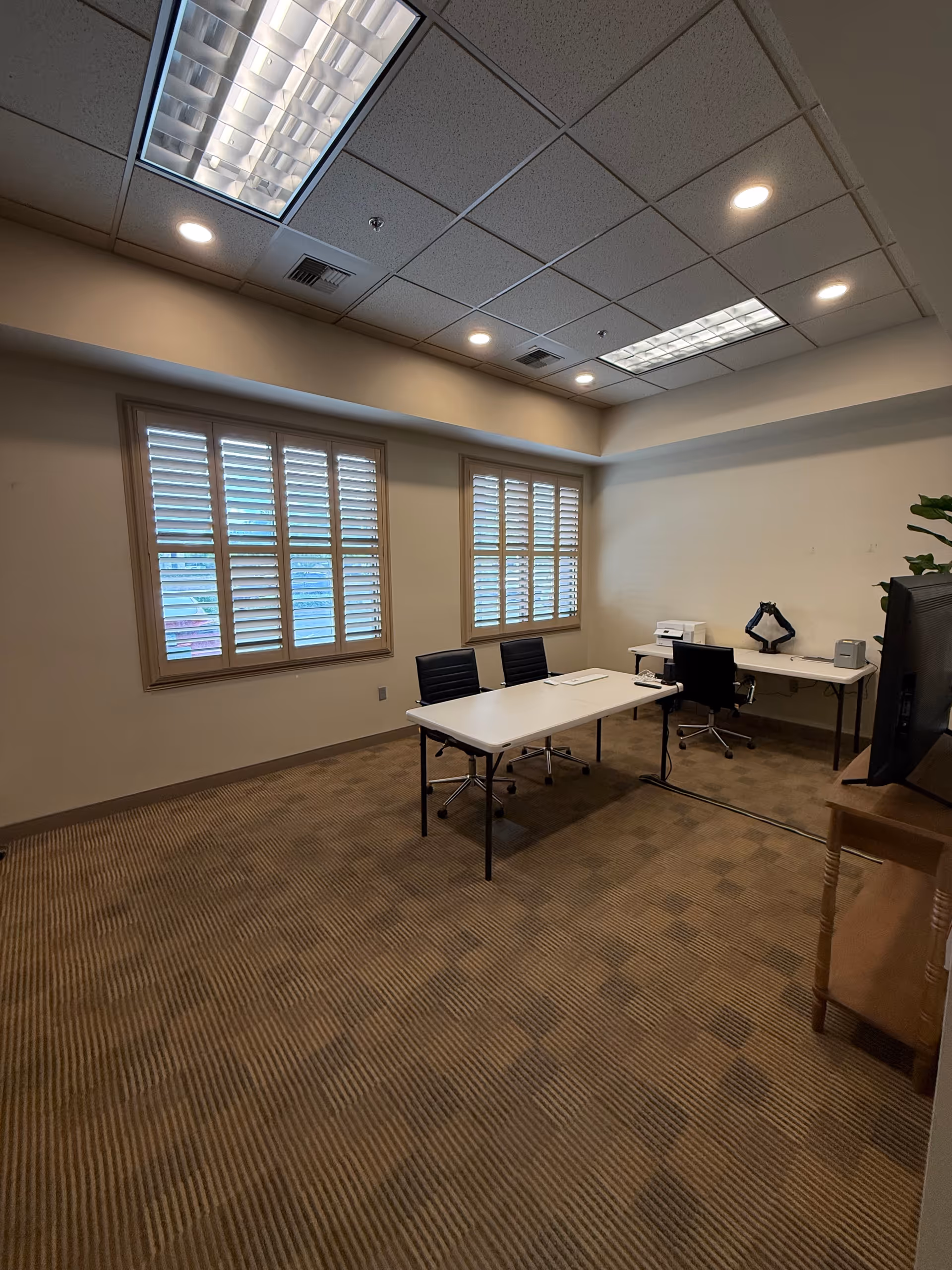 Sparse conference room with two desks, chairs, a printer, a monitor, and shuttered windows.