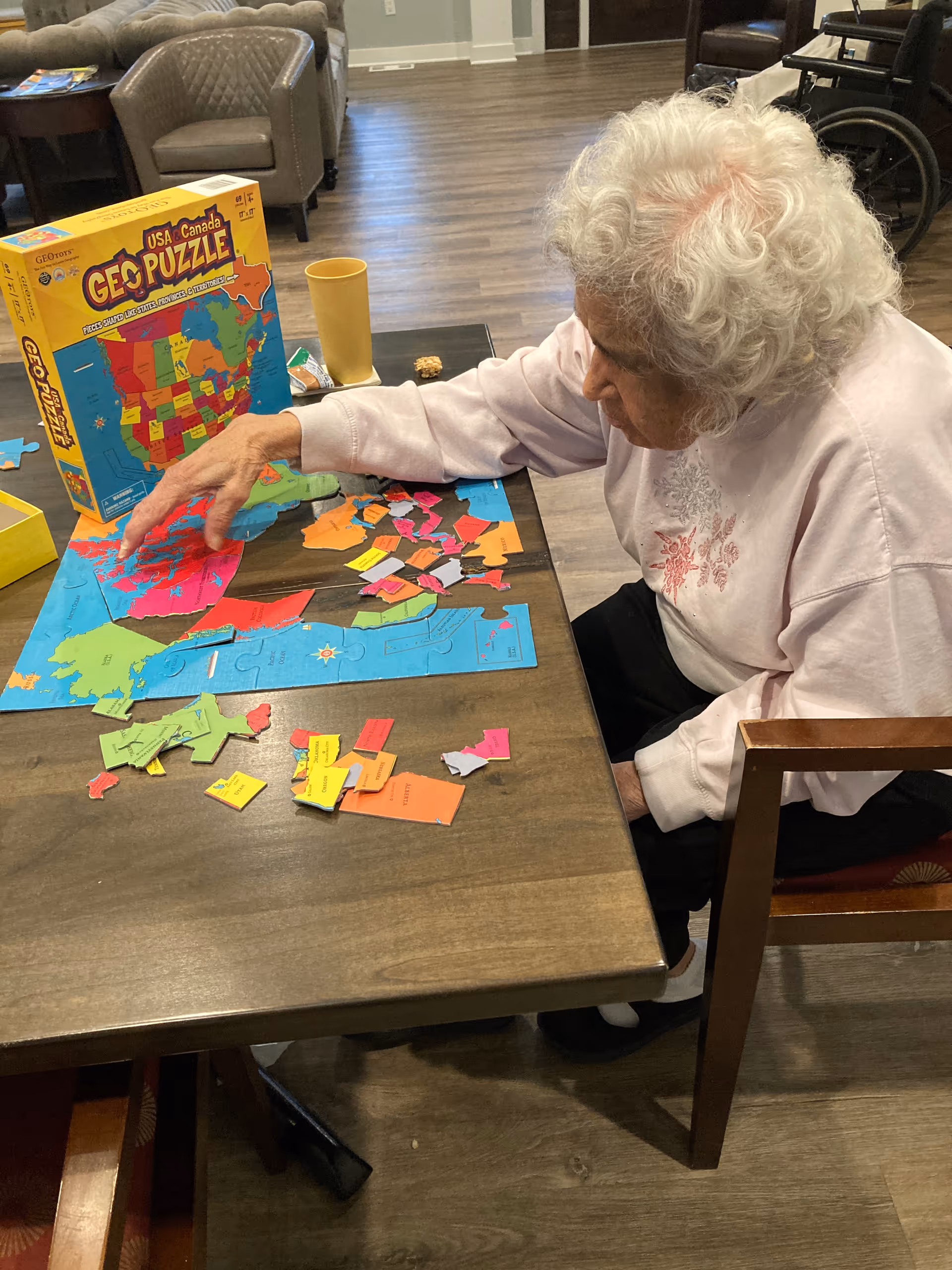 An elderly woman with white curly hair is sitting at a wooden table working on a colorful USA and Canada geo puzzle. The puzzle pieces are spread out on the table, and the puzzle box is standing upright nearby. The room has wooden flooring and comfortable chairs in the background.