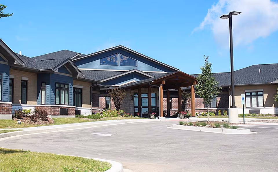 Exterior view of a single-story senior living facility building with a covered entrance, brick and siding facade, landscaped greenery, and a parking area under a clear blue sky.
