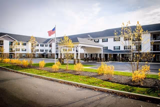 Exterior view of a large, three-story senior living facility with white siding and dark roofing. The building has multiple windows and a covered entrance with a driveway. There are small trees with yellow leaves and landscaped areas in front, along with an American flag on a flagpole near the entrance.