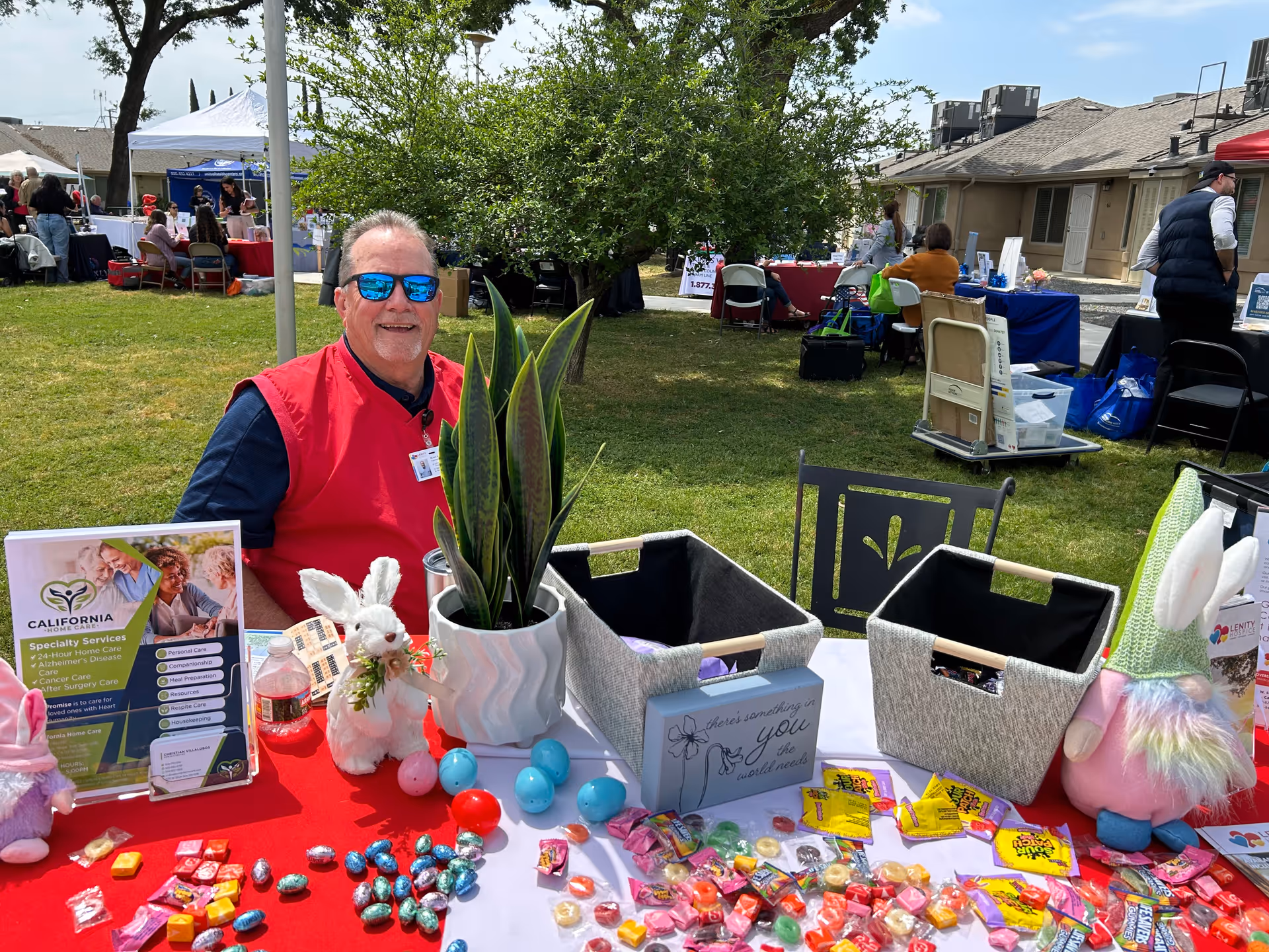 Smiling man in red vest sits at a booth with candy and plants outdoors.