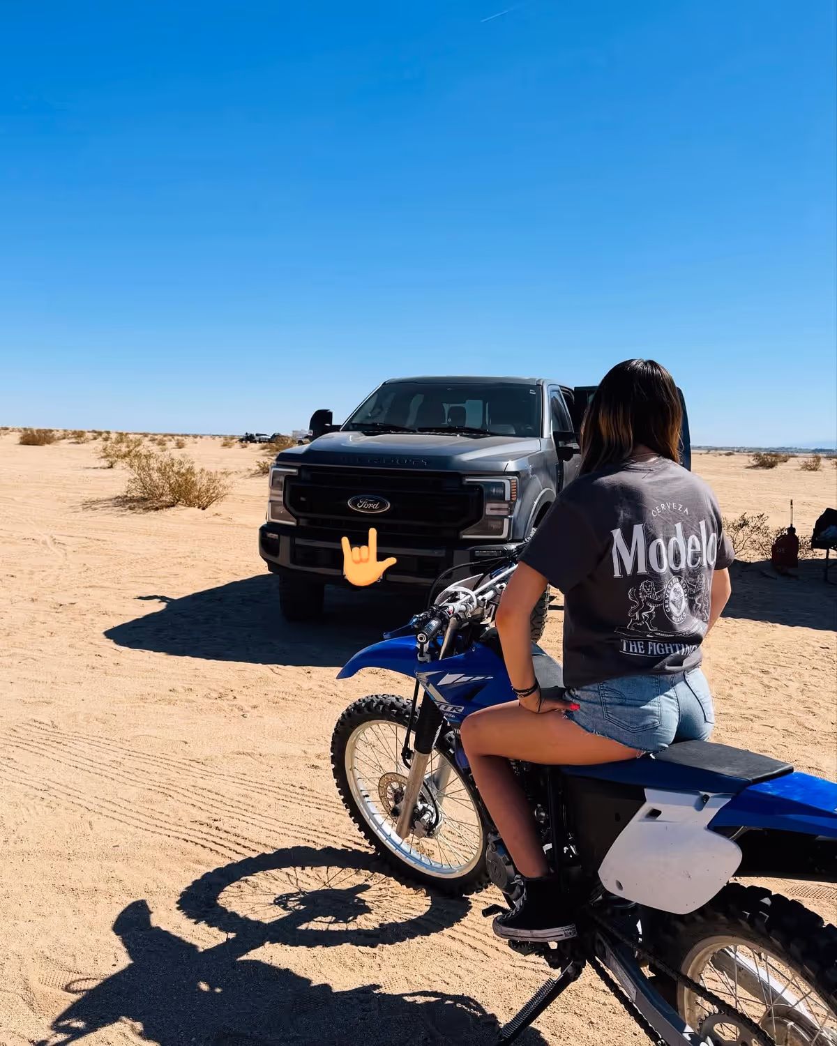 Person sits on a blue dirt bike near a gray Ford truck in a desert.