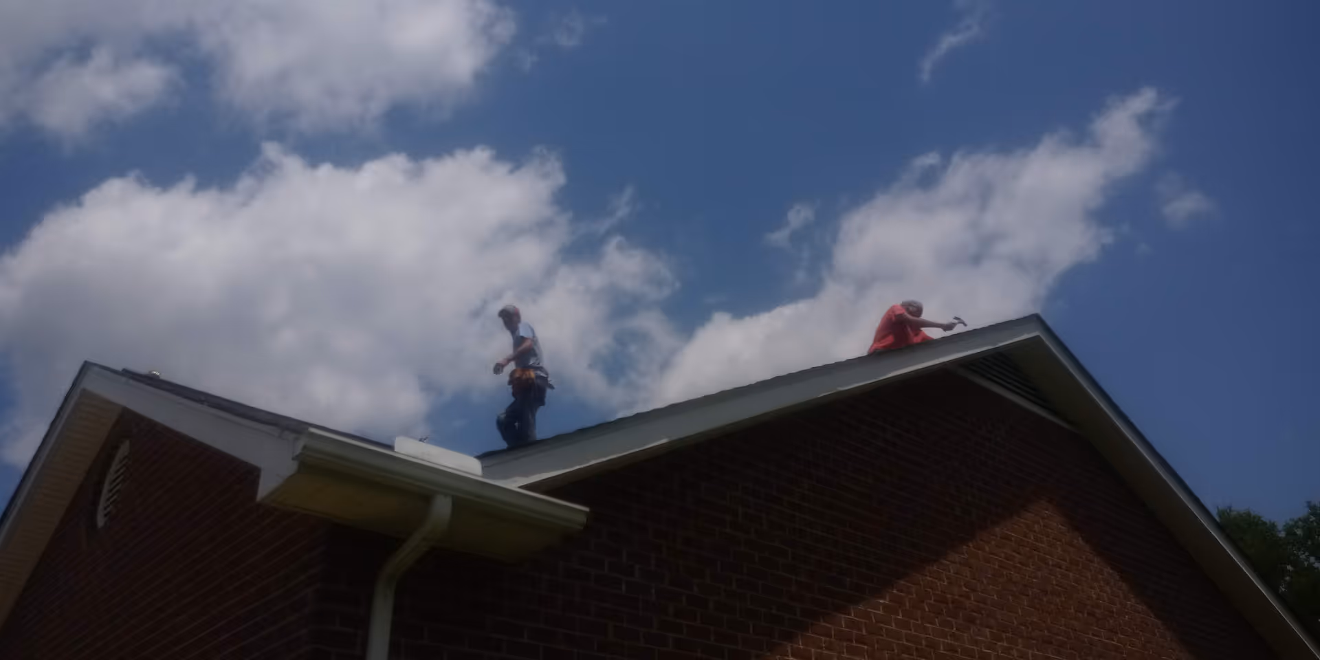 Two workers stand on the sloped roof of a brick building under a blue sky.