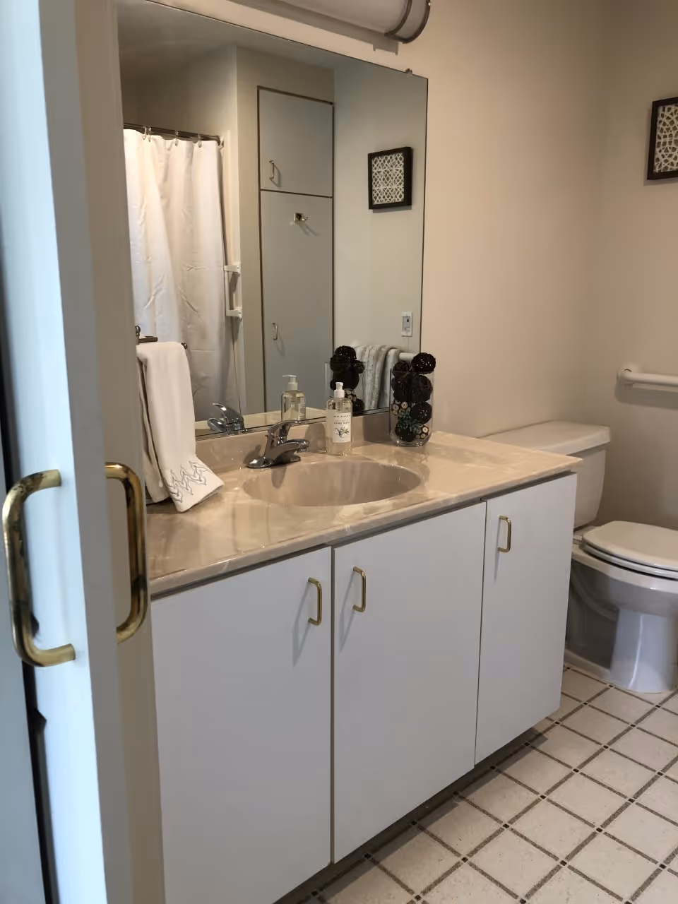 A clean bathroom with a beige countertop sink, a large mirror above the sink, a white cabinet with gold handles below, a toilet with a grab bar on the wall next to it, and a shower with a white curtain in the background.