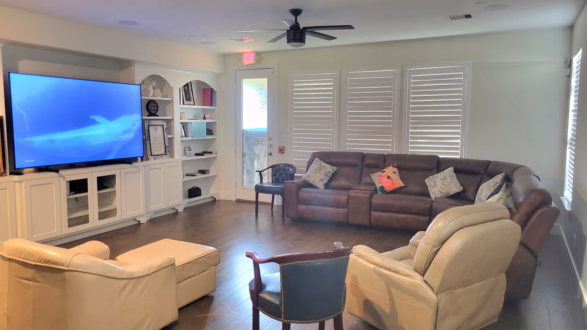 Bright living room with a large sectional sofa, recliners, TV, shelves, and windows.