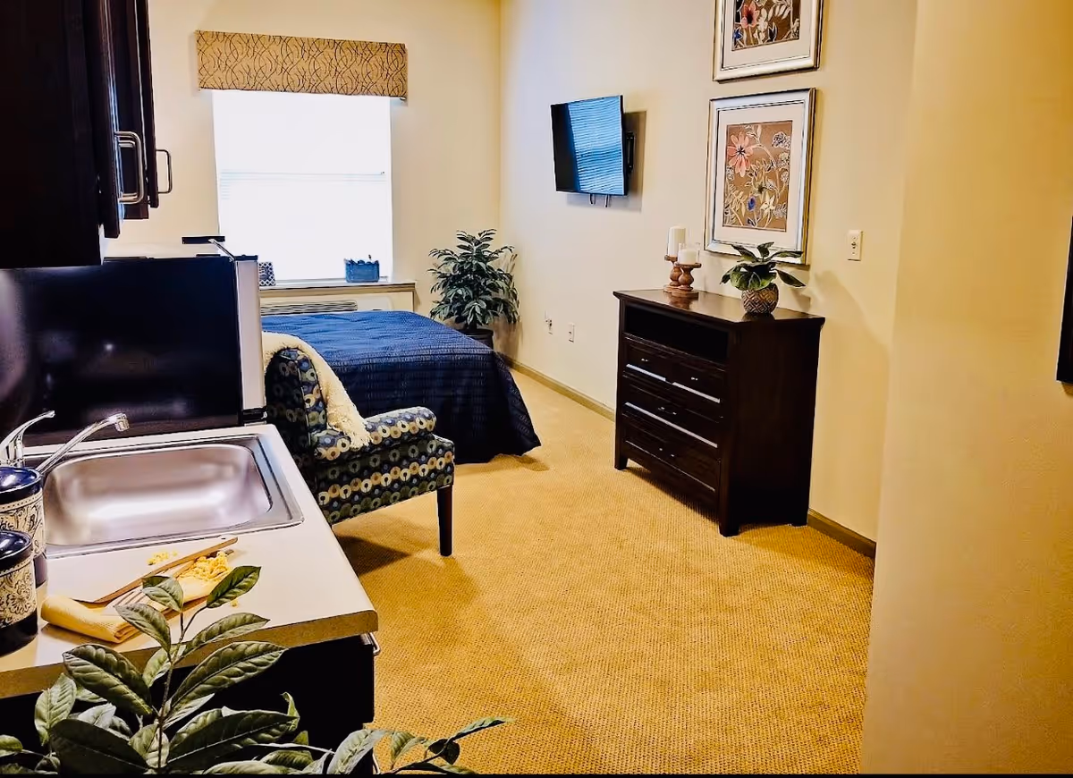 Interior view of a senior living facility room featuring a small kitchenette with a sink and counter, a patterned armchair, a bed with a dark blue bedspread near a window, a wall-mounted TV, a dark wooden dresser with decorative items, and framed floral artwork on the wall.