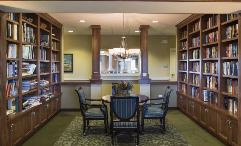 A cozy library room with wooden bookshelves filled with books on both sides. In the center, there is a round wooden table with four striped upholstered chairs around it. A chandelier hangs above the table, and a window with blinds is visible on the back wall.