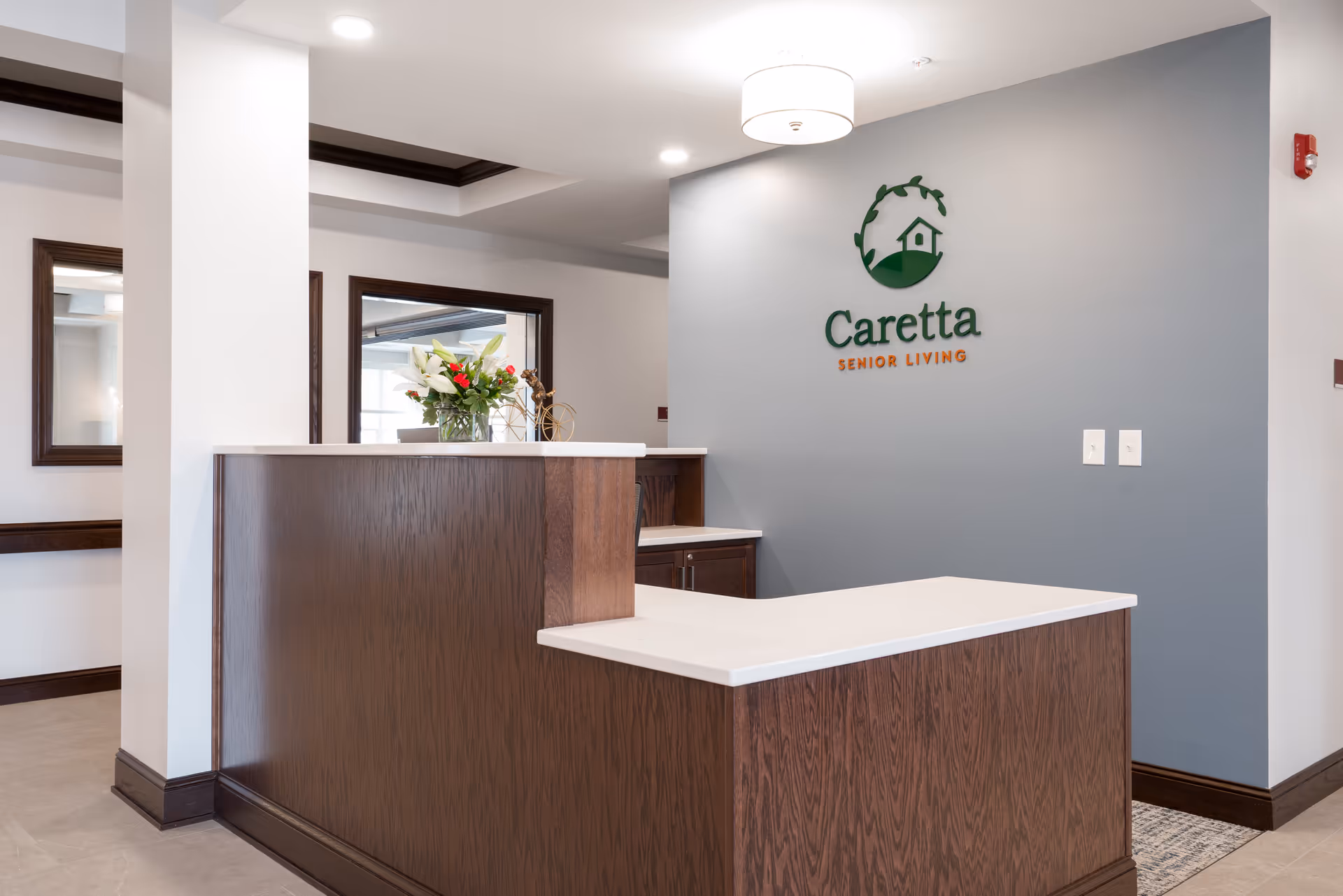 Reception desk with wood counters, a vase of flowers, and a 'Caretta Senior Living' logo on a gray wall.