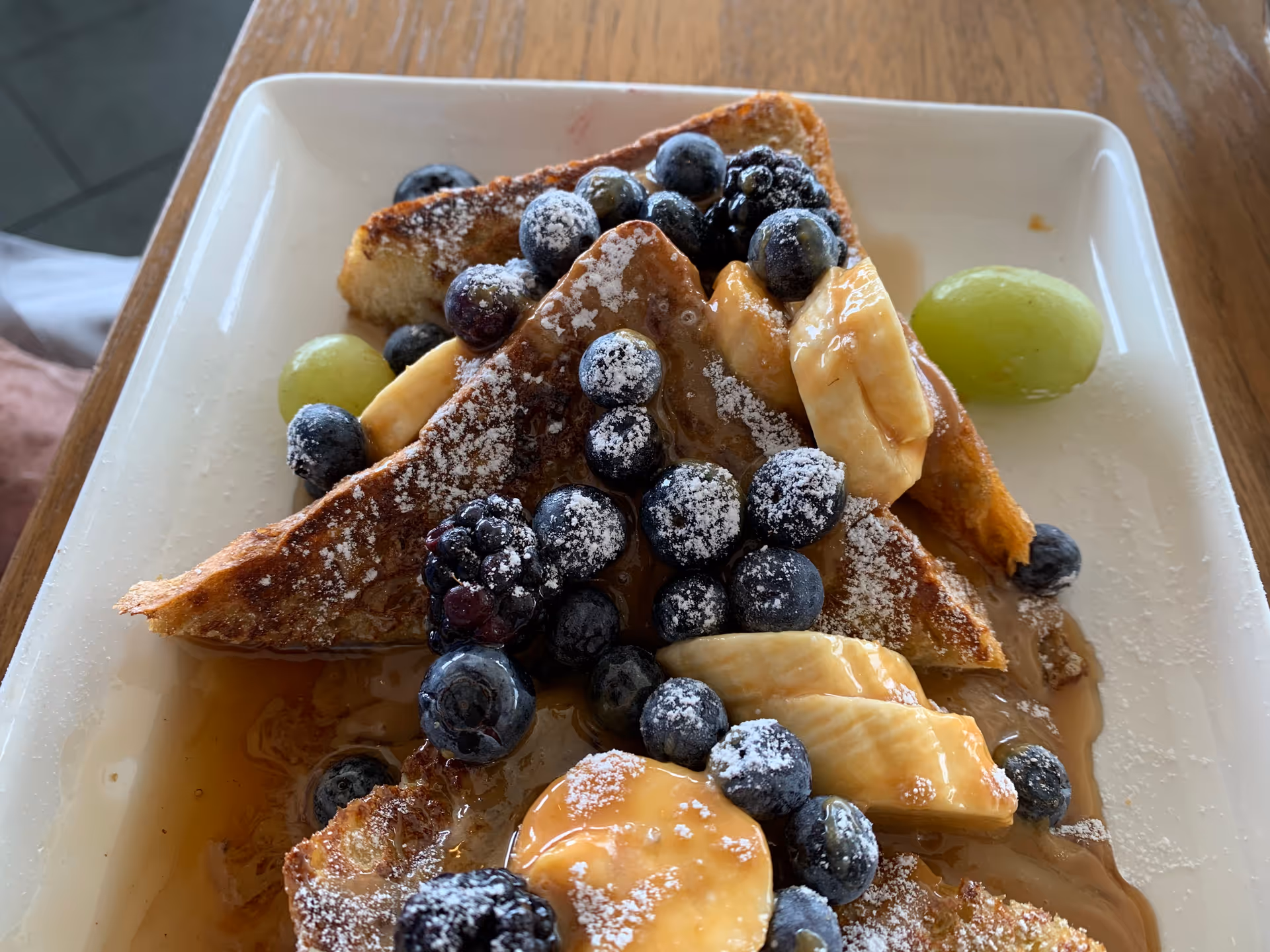 Close-up of French toast topped with blueberries, banana slices, powdered sugar, and syrup on a white plate.